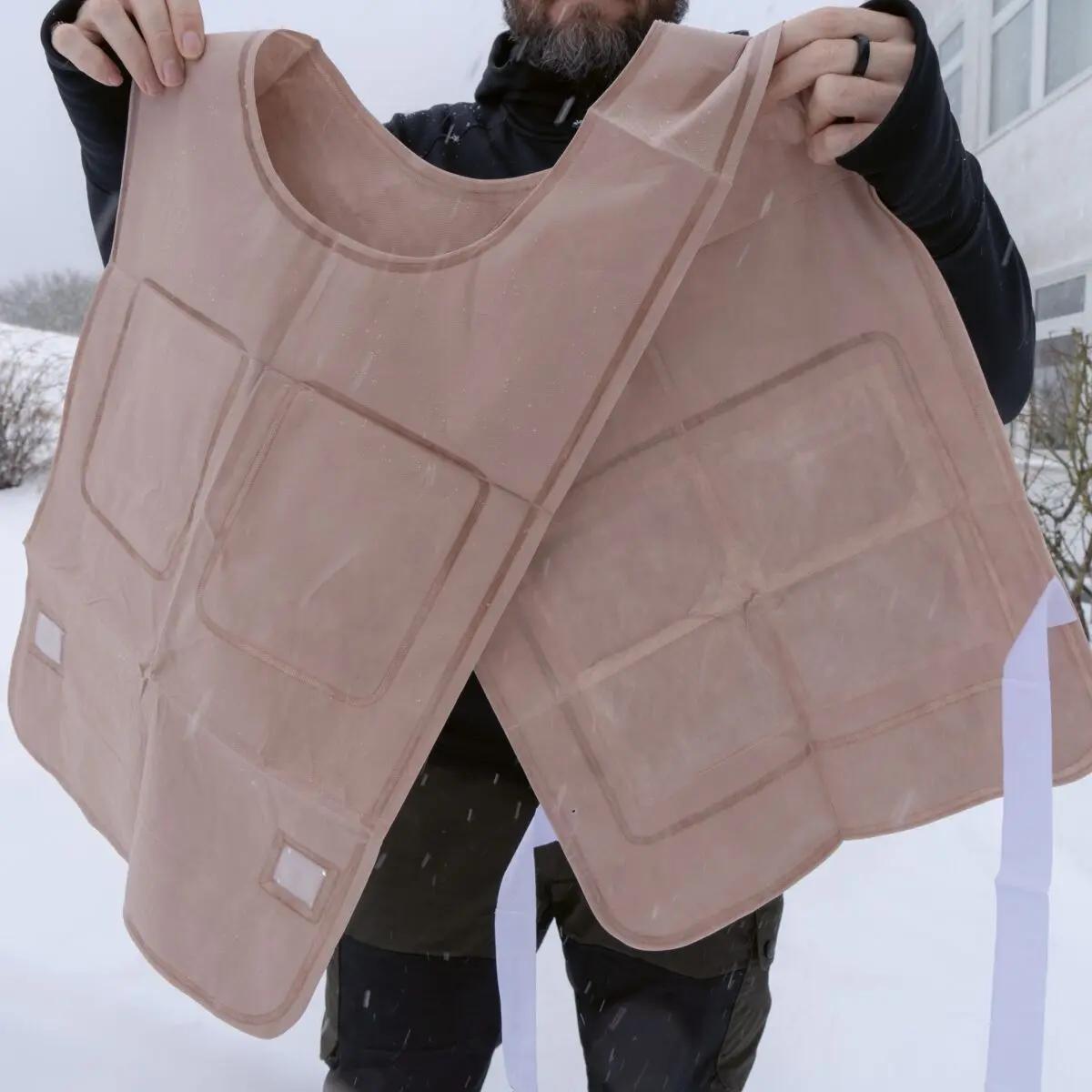 Eine Person hält zwei beige Ready-Heat™ - Weste aus wasserfester Kunstfaser mit mehreren Klettverschlusspaschen in winterlicher Schneelandschaft.