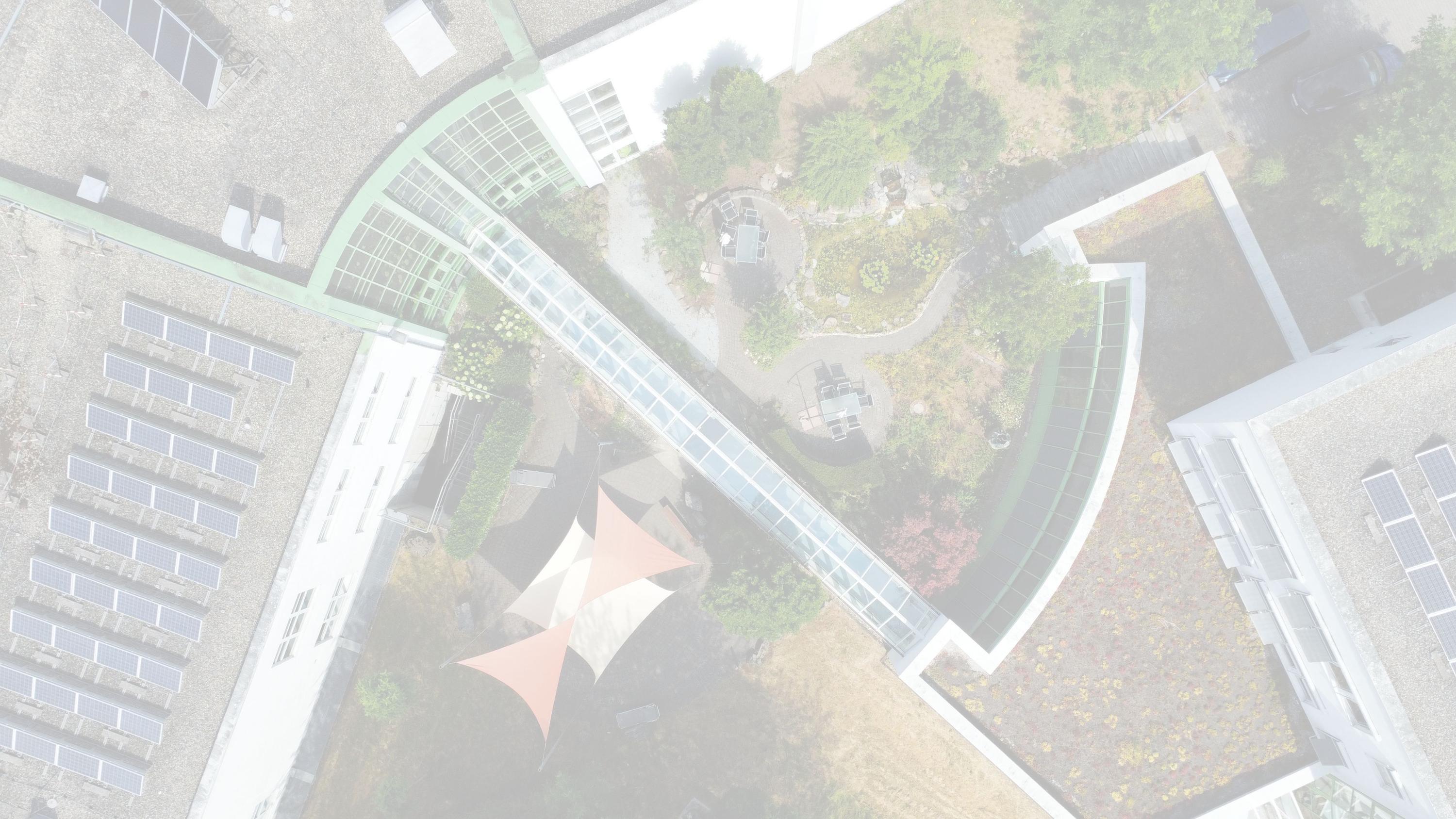 Moderne Gebäudestruktur mit gläserner Atriumbrücke, weißen Baukörpern, begrünten Flächen, geschwungenen Wegen und Parkplatz mit Autos.