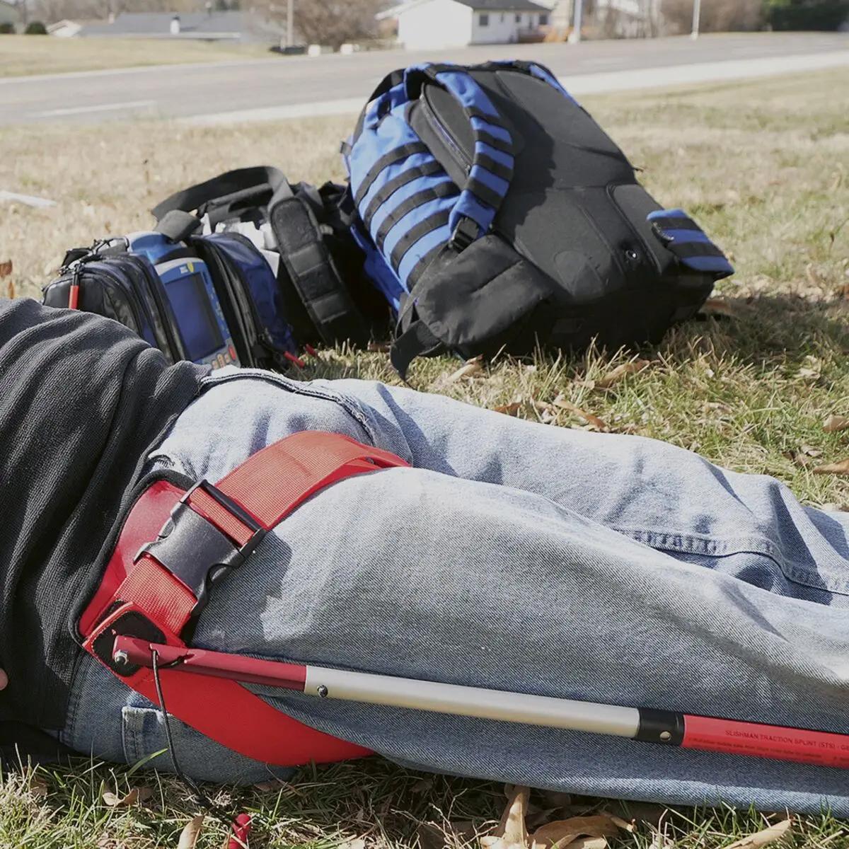 Eine Slishman Traction Splint (STS) GEN. 2 in rot-schwarz liegt neben einer Person auf der Wiese, dazu gehört ein roter Hüftgurt und weiteres Notfallequipment.