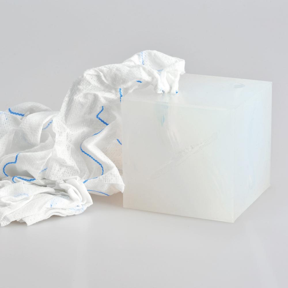Ein transparenter, weiß-blauer Wound Cube™ Schwamm liegt neben einer weißen, strukturierten Kunststoffbox auf einer hellgrauen Fläche.