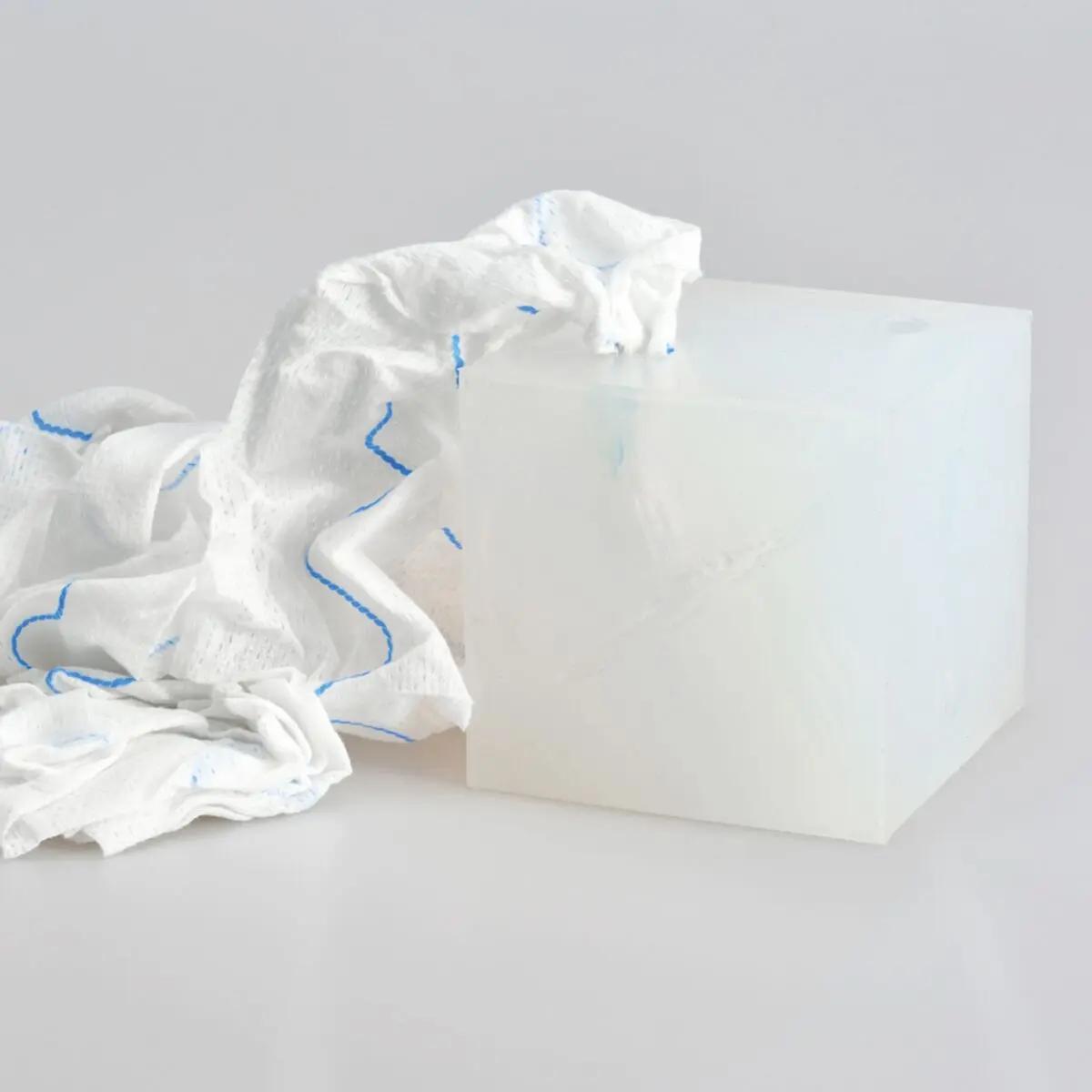 Ein transparenter, weiß-blauer Wound Cube™ Schwamm liegt neben einer weißen, strukturierten Kunststoffbox auf einer hellgrauen Fläche.