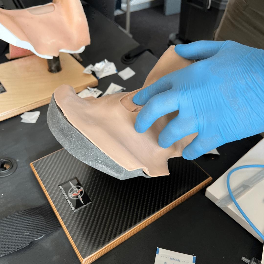 Eine Hand mit blauem Handschuh hält einen Battlefield Partners Desktop CRIC Trainer aus beigefarbenem Material auf einer schwarzen Carbonfaser-Basis mit Logo.