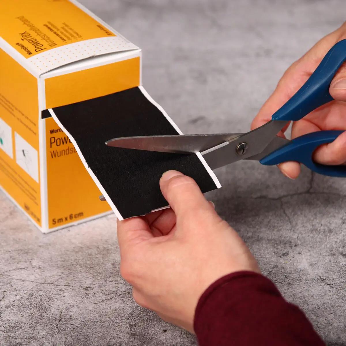 Eine Hand schneidet ein schwarzes Stück Wundschnellverband Weroplast® PowerTex - 6 cm, 5 m mit blauen Scheren von einer gelben Verpackung ab.