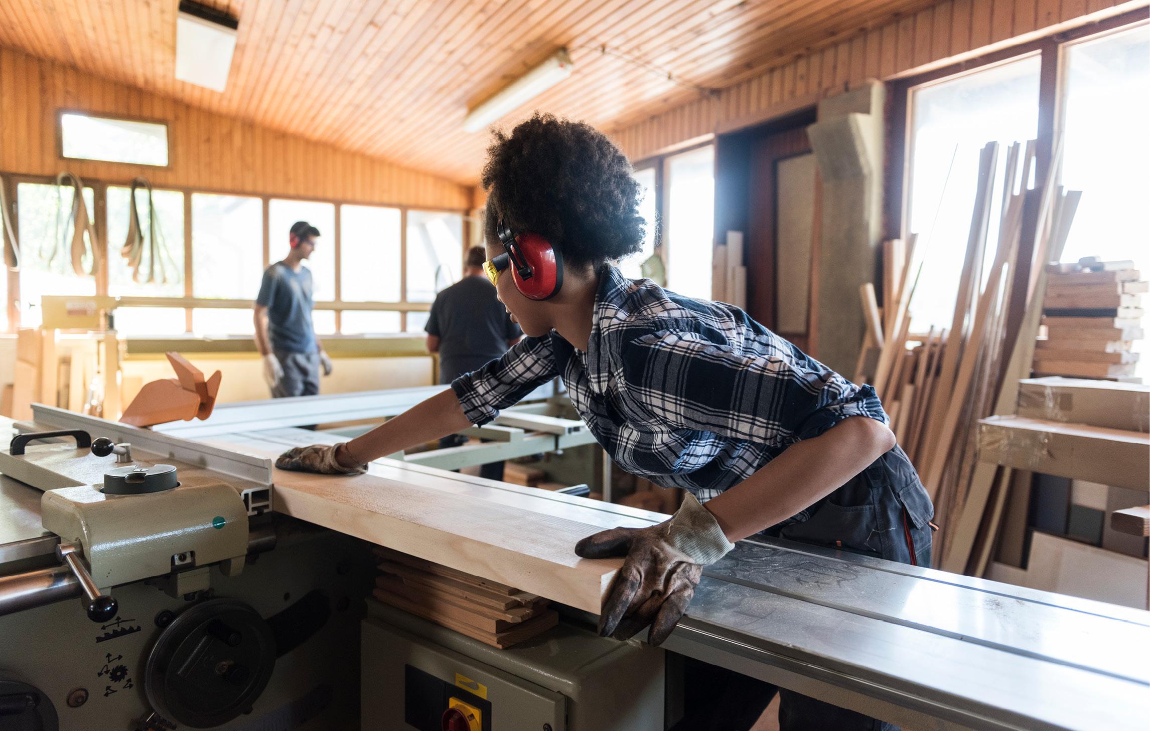 Alltagssituation in einer Schreinerei. Eine Frau trägt Gehörschutz und durchsägt an einer automatischen Sägemaschine eine Holzlatte