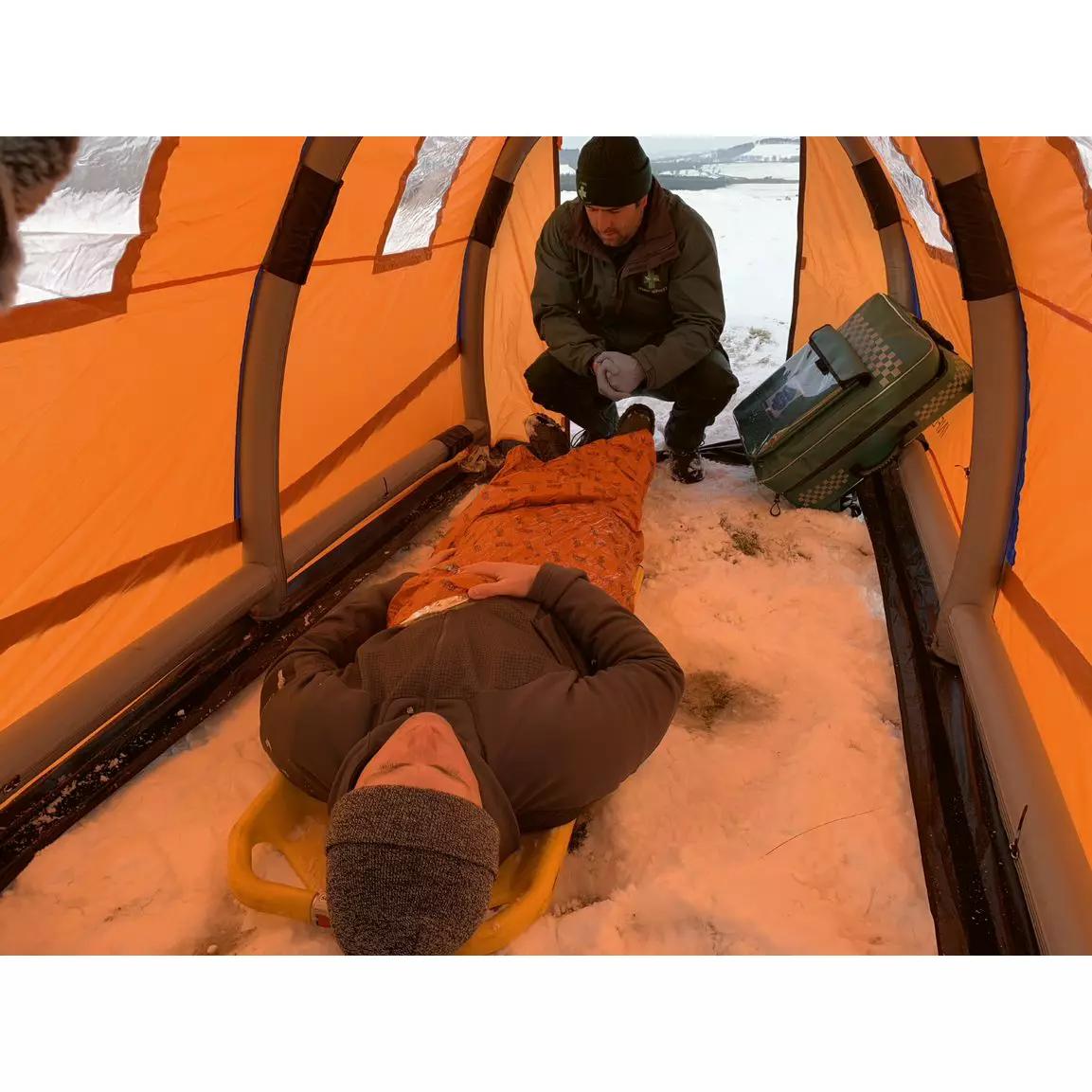 Ein Mann liegt auf einer Trage im RIGLOO Rescuer Rettungszelt, das leicht, sturmsicher und zum Schutz bei Versorgung vor Ort aufgeblasen ist.