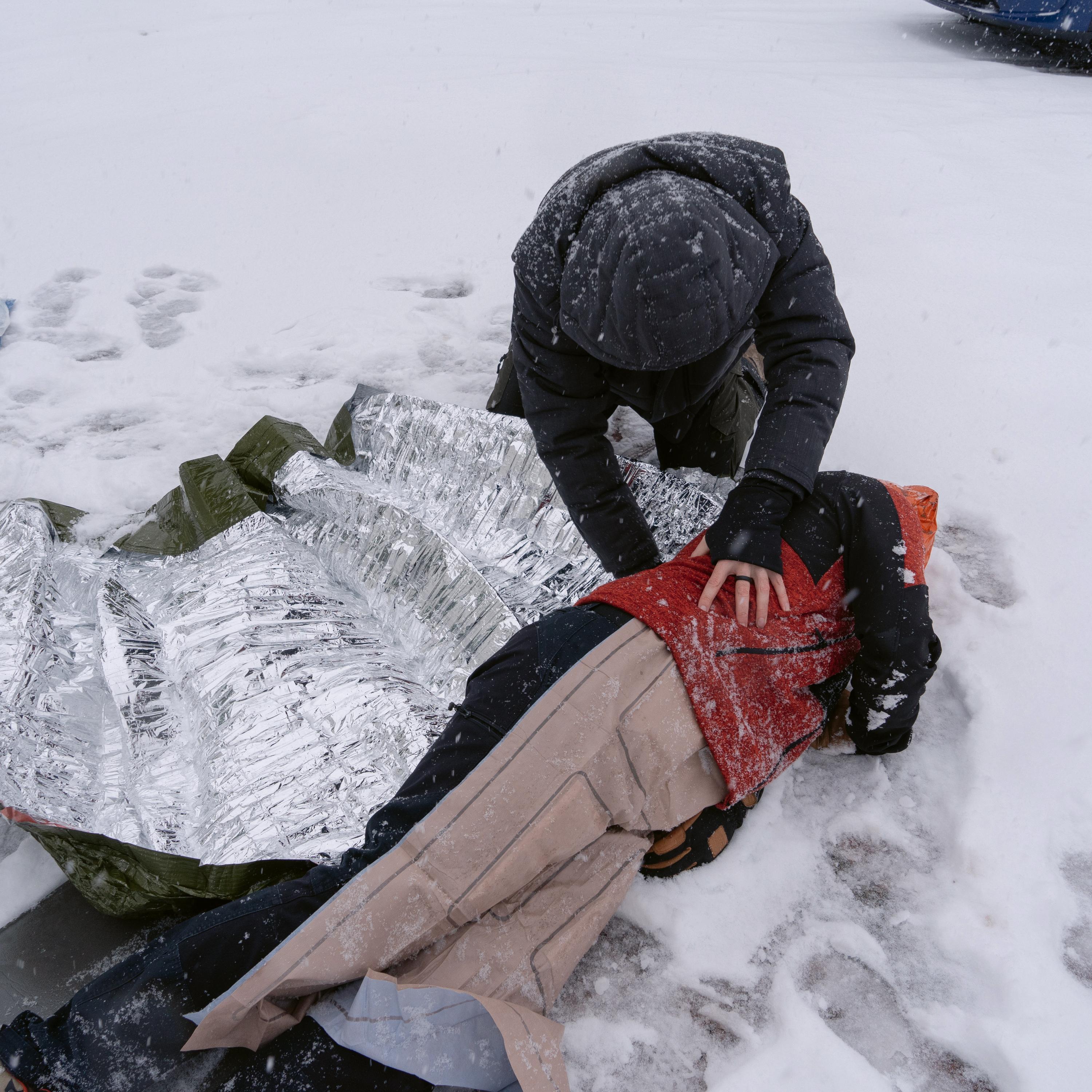 Eine Person legt im Schnee eine Blizzard - Grün, Decke schützend um eine verletzte Person, deren Körper von einer Eisschicht bedeckt ist.