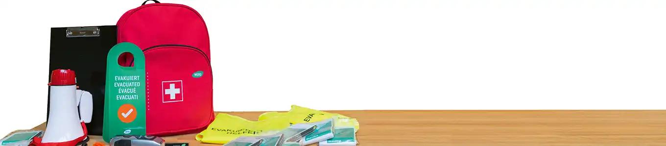 Rotes Erste-Hilfe-Set mit Megafon, Klemmbrett, grünem Ständer mit Häkchen sowie gelben und grünen Notfallbeuteln auf Holztisch