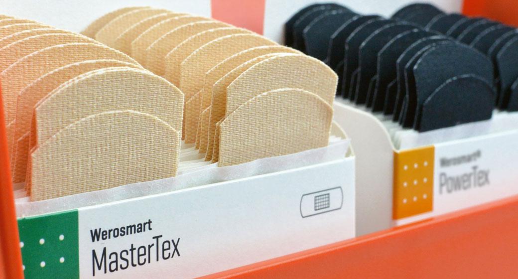 Rote Aufbewahrungsbox mit zwei Kisten: links helle, textile Werasmart® MasterTex Pflaster, rechts schwarze, runde Werasmart® PowerTex Pflaster.