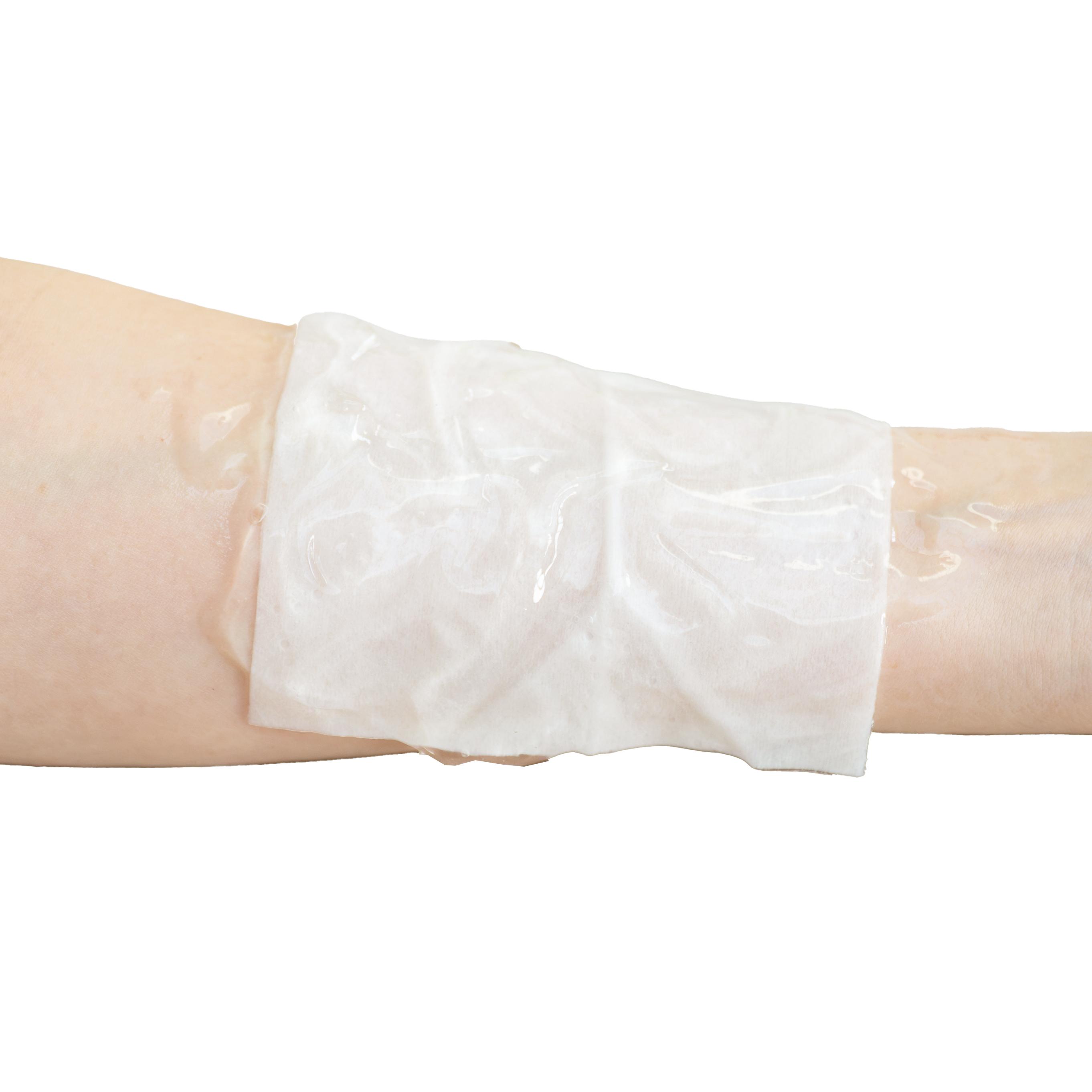 WERO BurnAway® Verbrennungskompresse, 10 x 10 cm, mit leicht strukturierter, durchscheinender Oberfläche auf einen Arm gewickelt, transparenter Flüssigkeitströpfchen daneben