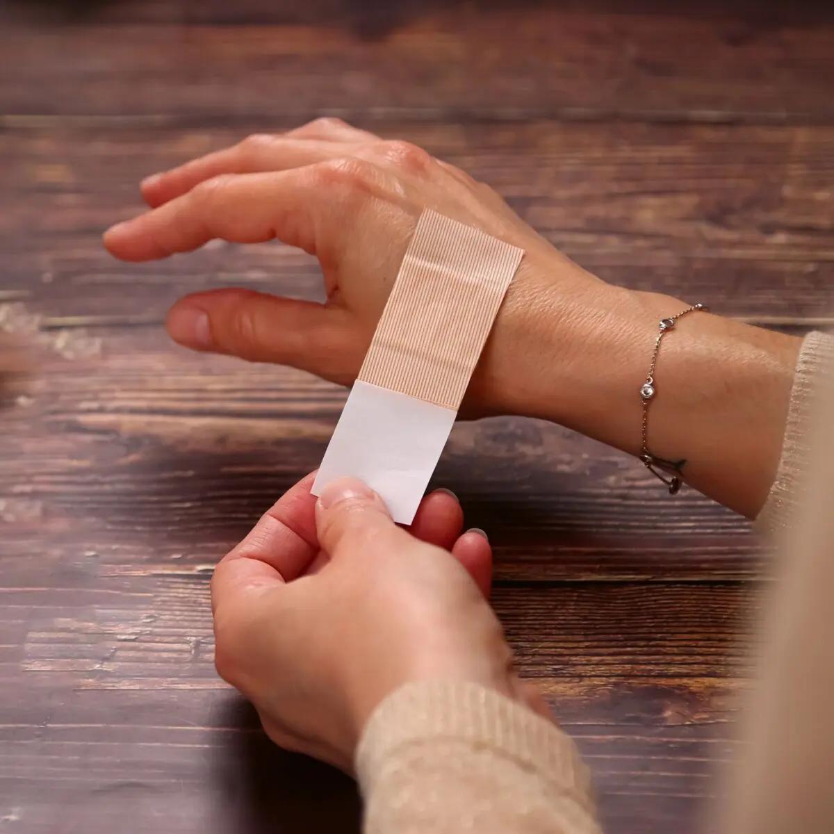 Eine Hand mit rot lackierten Fingernägeln und silbernem Armband trägt ein beiges Pflasterset Weroplast® AquaFlex DIN 13157, das um das Handgelenk geklebt ist.