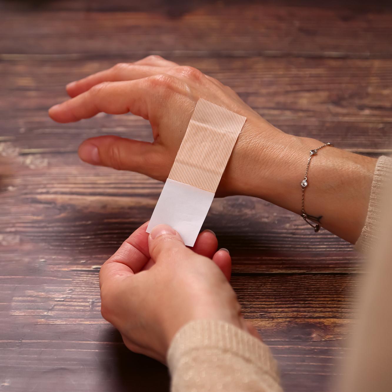 Eine Hand mit rot lackierten Fingernägeln und silbernem Armband trägt ein beiges Pflasterset Weroplast® AquaFlex DIN 13157, das um das Handgelenk geklebt ist.
