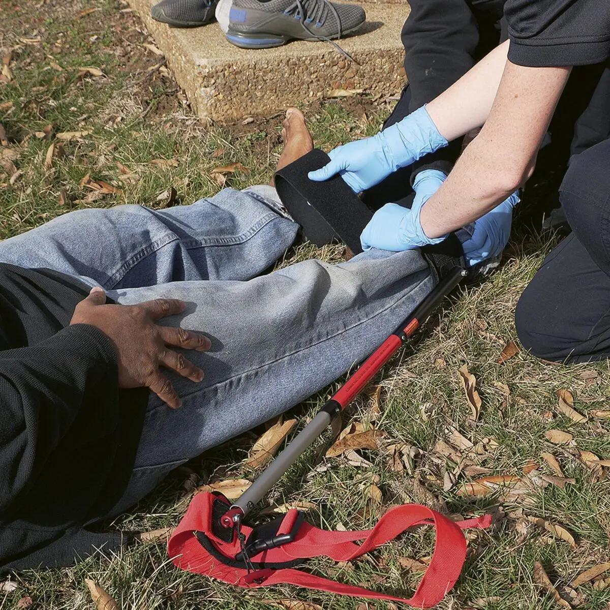 Eine verletzte Person erhält mithilfe der Slishman Traction Splint (STS) GEN. 2 eine schwarze Traktionsschiene am Bein, während zwei Helfer Erste Hilfe leisten.