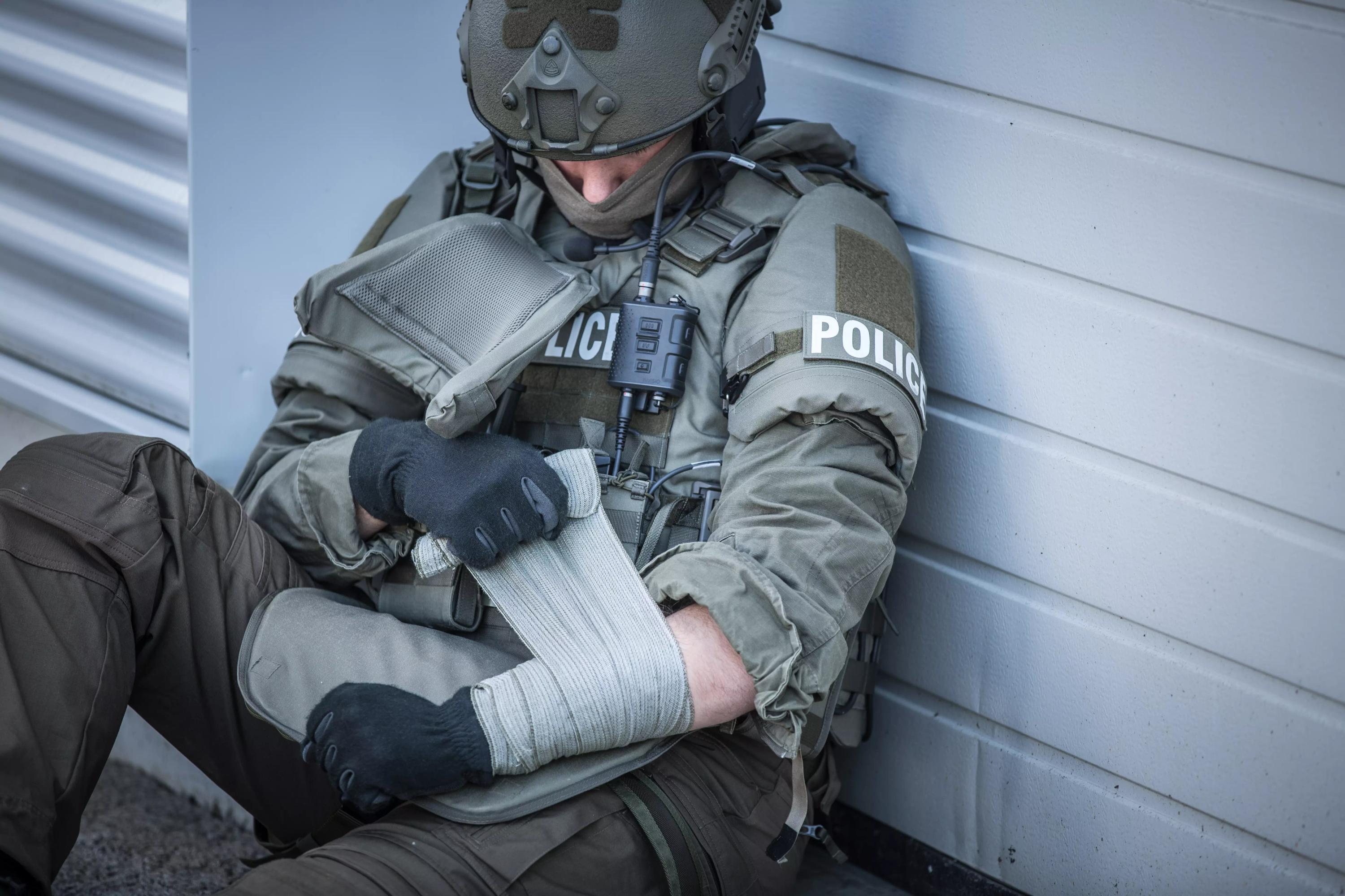 Eine Person im grauen Polizeiuniform legt einer verletzten Kollegin mit der Emergency Bandage®, das Original - Oliv, 15 cm einen Verband am Handgelenk an.
