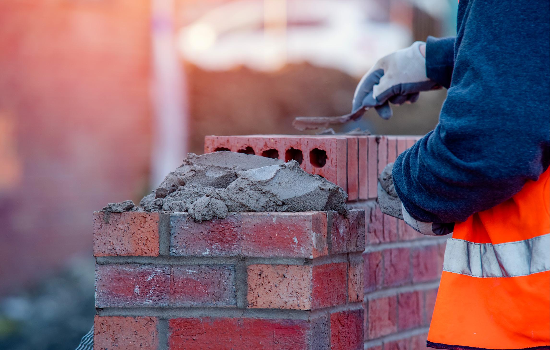 Person mit Warnweste und Arbeitshandschuhe zementiert eine Mauer