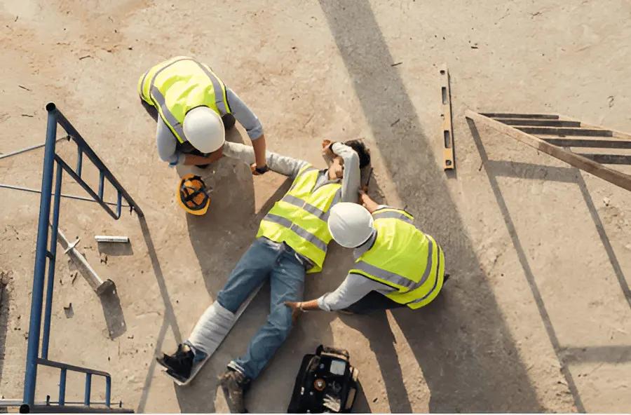 Zwei Bauarbeiter mit gelben Warnwesten, einer verarztet den anderen mit bandagiertem Bein, umgeben von Werkzeugen auf einer Baustelle.