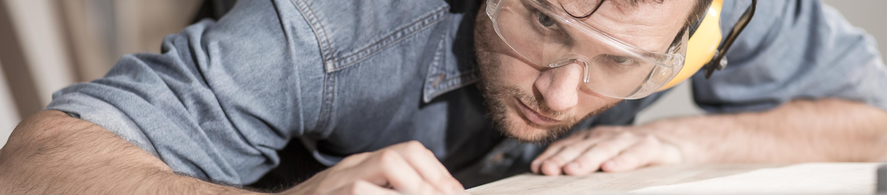 Eine Person prüft mit den Augen und Händen eine Holzplanke und trägt dabei eine Schutzbrille.