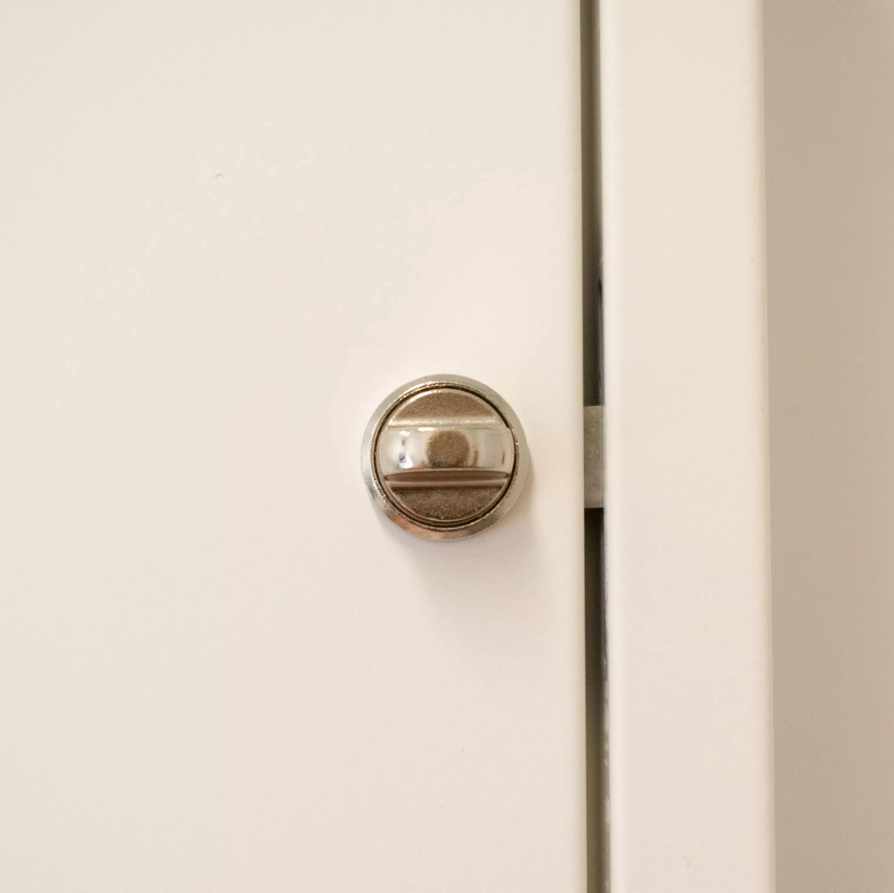 Nahaufnahme eines silbernen Ersatz-Drehknopfes für WERO Verbandschränke mit mittigem Grat und zwei Metallbändern auf weißer Tür.