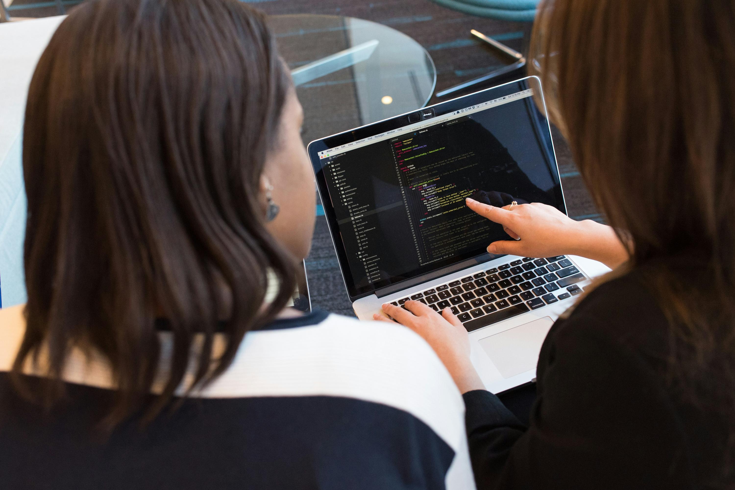 Zwei Frauen sehen sich gemeinsam Code auf einem Laptop an, eine navigiert mit der Hand auf dem Trackpad.