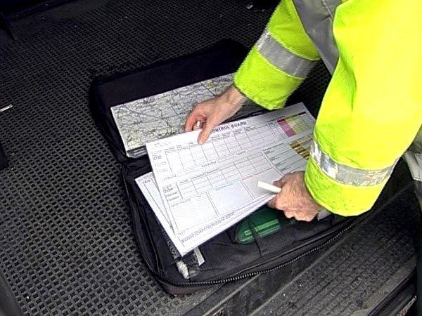 Person in gelber Warnjacke prüft konzentriert ein Klemmbrett mit technischem Kontrollboard und Karte auf einem schwarzen Koffer.