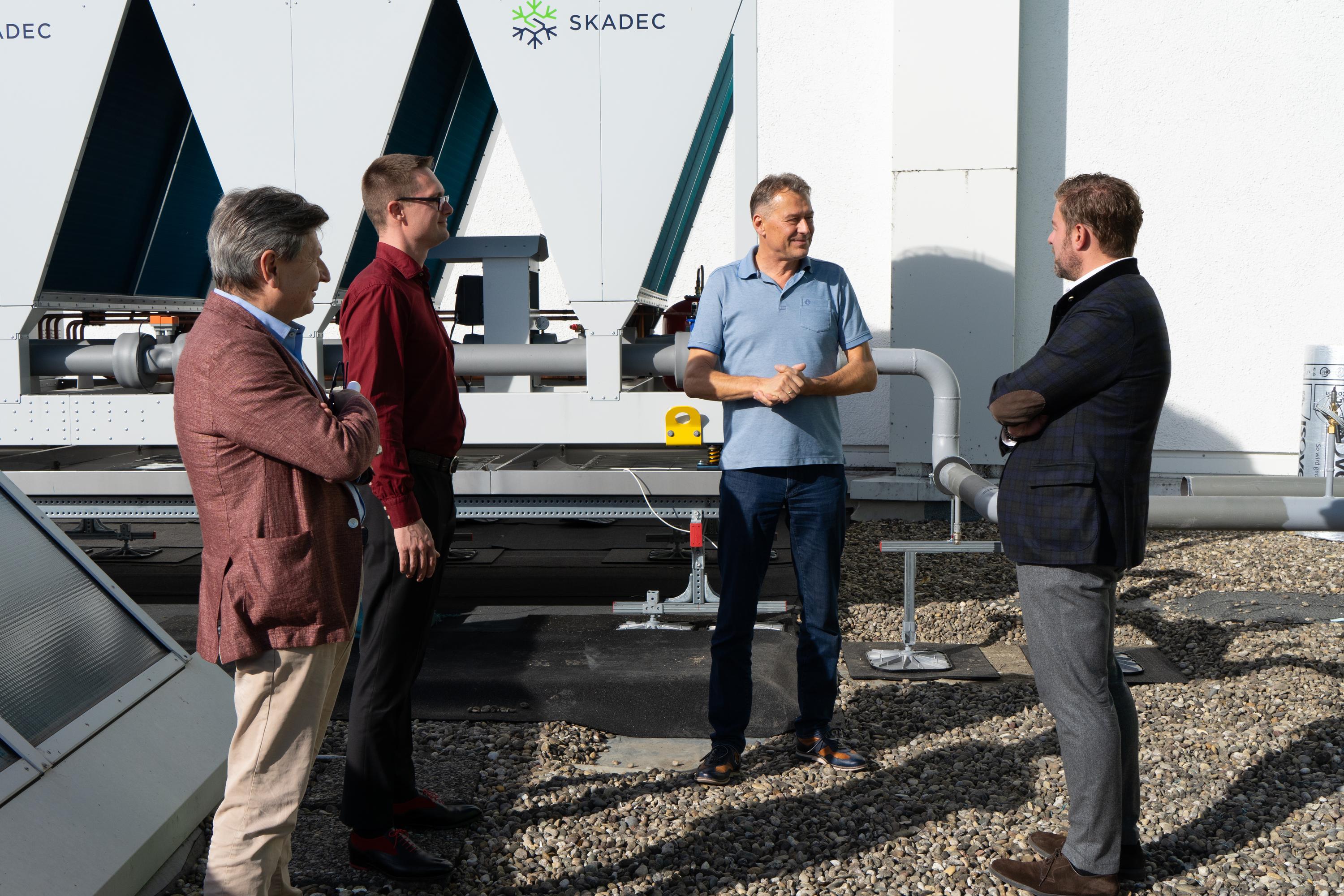 Vier Männer unterhalten sich vor einem weißen SKADEC-Anlagenmodul mit turquoisefarbenen Rohren auf einem Kiesdach.