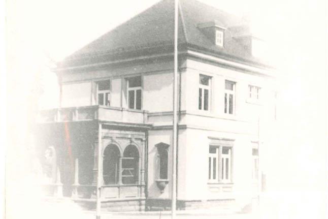 Zweistöckiges Haus mit dunklem Dach, heller Stein- oder Ziegelfassade, mehreren Fenstern und einer Veranda mit Rundbögen.