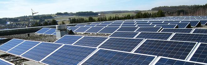 Dunkelblaue Solarpaneele in Reihen auf einem Dach, daneben ein kleines Gebäude, im Hintergrund grüne Landschaft mit Bäumen und Kran.